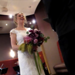 A bride in white lace gown with bouquet of mixed purple flowers laughs during her weddng ceremony. Photo by BF Newhall