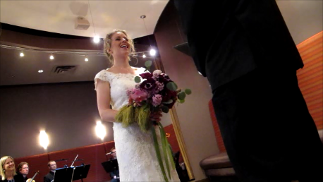A bride in white lace gown with bouquet of mixed purple flowers laughs during her weddng ceremony. Photo by BF Newhall