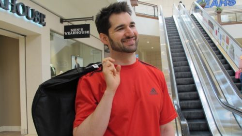 A relaxed groom in red t-shirt smiles and carries his Mens's Wearhouse tux in a bag. Photo by BF Newhall