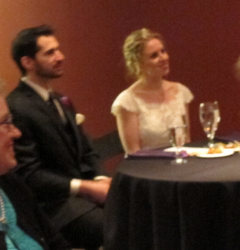 Brideand groom seated at a table with black tablecloths being toasted and smiling. Photo by BF Newhall