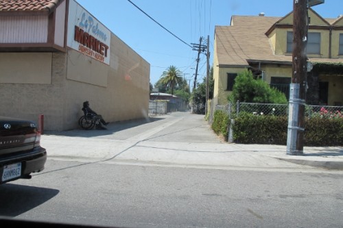 a man in a wheelchair sits in the shade of two-story building in a paved alley in los angeles. photo by bf newhall