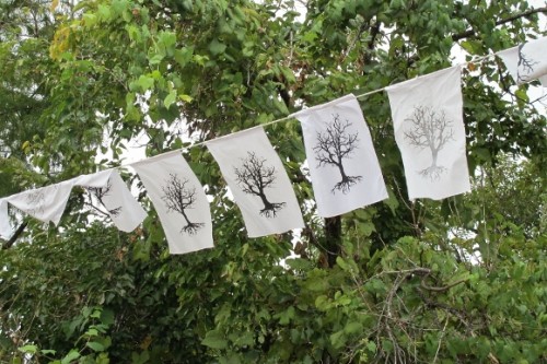 White prayer flags silk screened with images of dead trees hang in front of living green trees in Austin, TX. Photo by BF Newhall