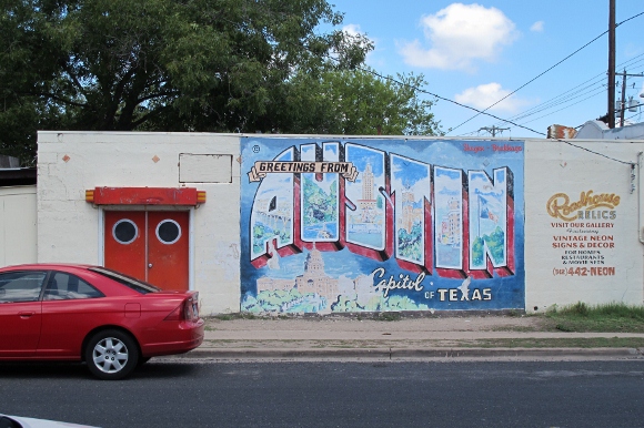 A blue and red graffiti mural on a wall in Austin TX shows a postcard reading "Greetings from Austin, capitol of Texas." Photo by BF Newhall