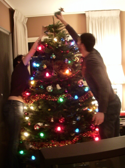 Two young adults place the angel atop a green Christmas tree decorated with lights. Photo by BF Newhall