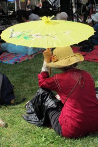 A woman in a red shirt sits in the sun at Berkeley's Himalayan Fair holding a yellow parasol over her head. Photo by BF Newhall