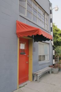The entrance to the gray Berkeley Potters Guild building is a red door with red awning. Photo by BF Newhall