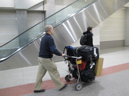 The politics of housework. A man pushes a cart stacked high with luggage at an airport. Photo by Barbara Newhall